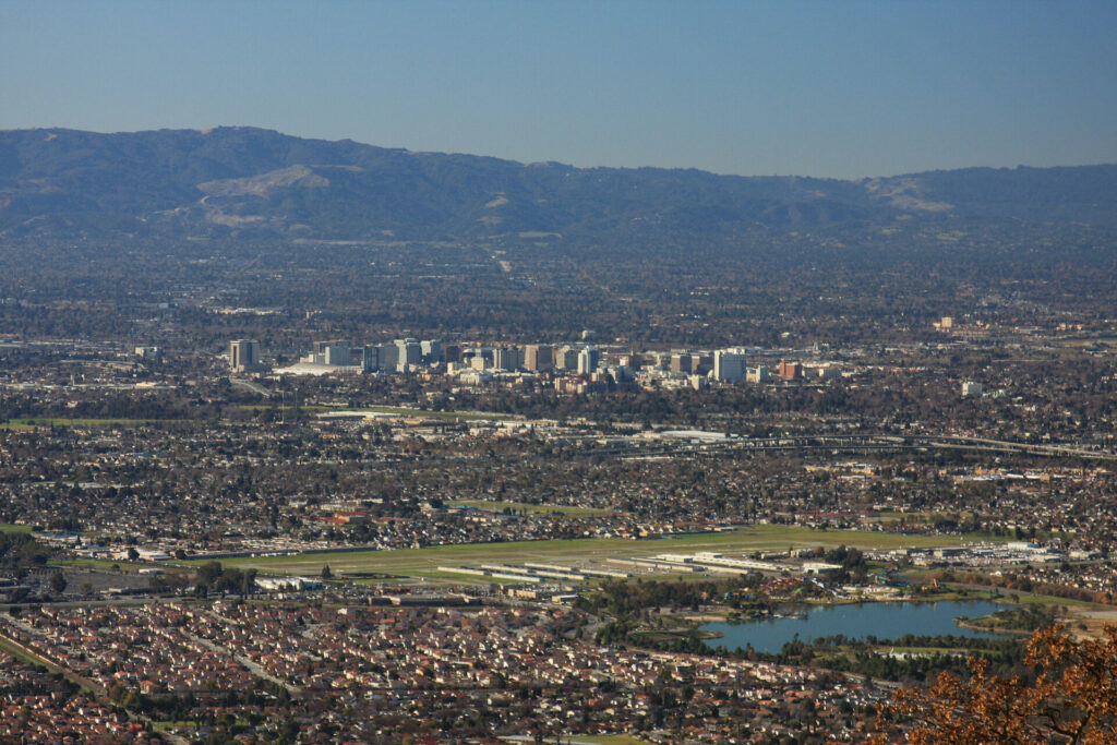 Aerial view of a suburban area with residential neighborhoods in the foreground, a mobile pet grooming van parked on a street, tall buildings in the center, and mountains in the background under a clear sky.