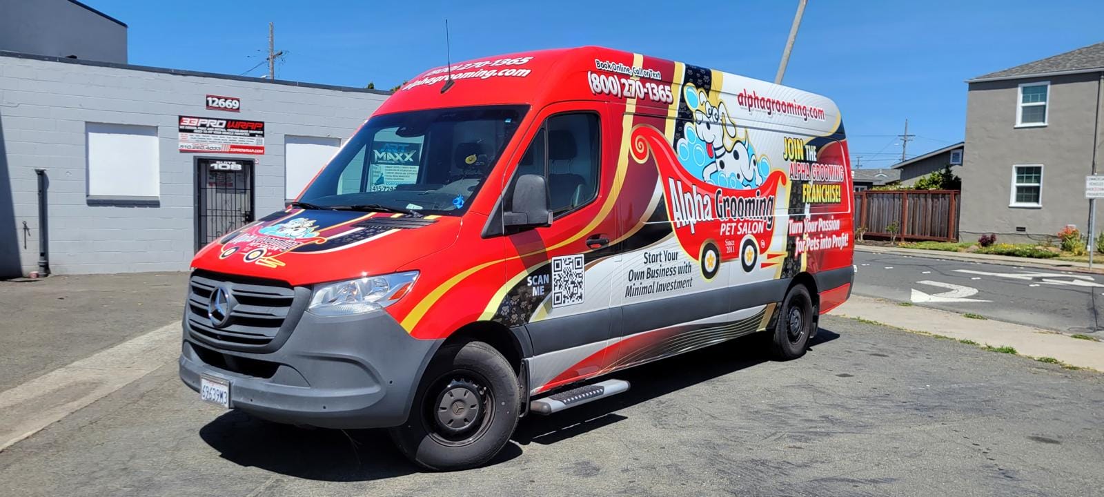 A red and white van with "Alpha Grooming Pet Salon" branding, highlighting mobile pet grooming, is parked on a street near a gray commercial building and a fenced yard.