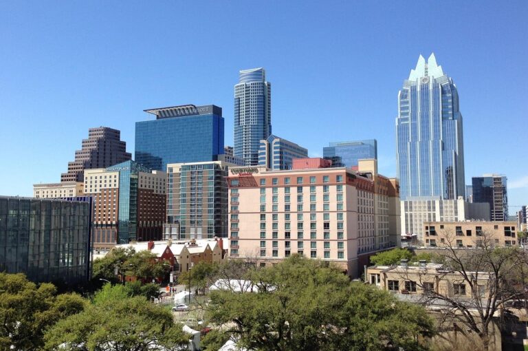 Downtown city skyline with modern skyscrapers, clear blue sky, and trees in the foreground.